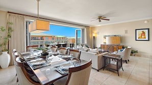 A spacious dining area with a large table set for a meal, a seating area with sofas and chairs, a ceiling fan, and a view of buildings through large windows. - Maui Resort Rentals: Honua Kai - Premier 7th Floor Penthouse, 4BR w/ Stunning Ocean and Black Rock Views! (Lahaina)