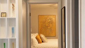 A narrow hallway leading to a bedroom with a bed, a white shelf with decorative items, and a framed picture on the wall. - Ultra Clean, Private, Comfy Home in Prime Location. Sleeps up to 8. (Milwaukee)