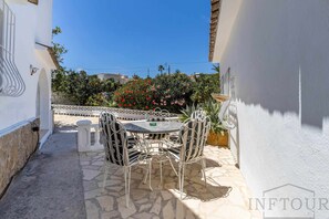 A patio with a round table and chairs, a white wall, and a view of a garden with red flowers. - Inftour La Fustera Villa (Benissa)