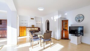 A spacious living area with a dining table, chairs, a TV, and a clock on the wall.