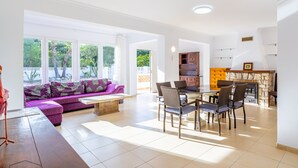 A spacious living area with a sofa, dining table, and chairs. There is a fireplace and a kitchen area visible in the background.