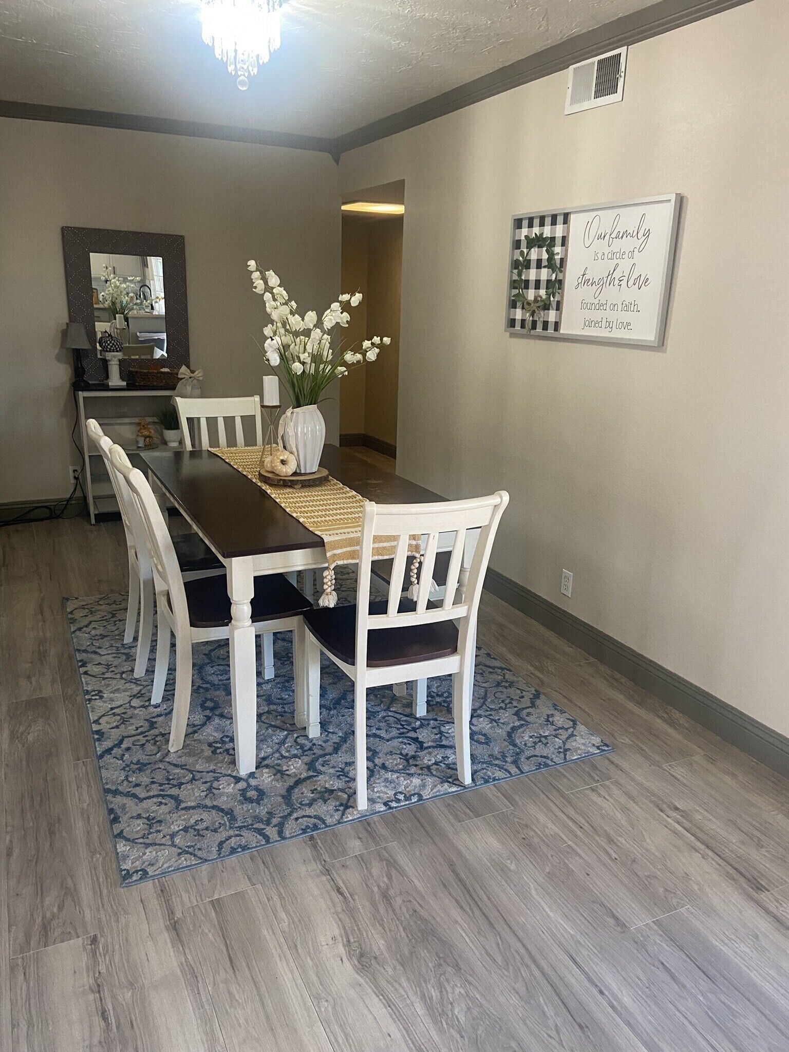 A dining area with a long table, white chairs, a vase with flowers, and a framed quote on the wall.