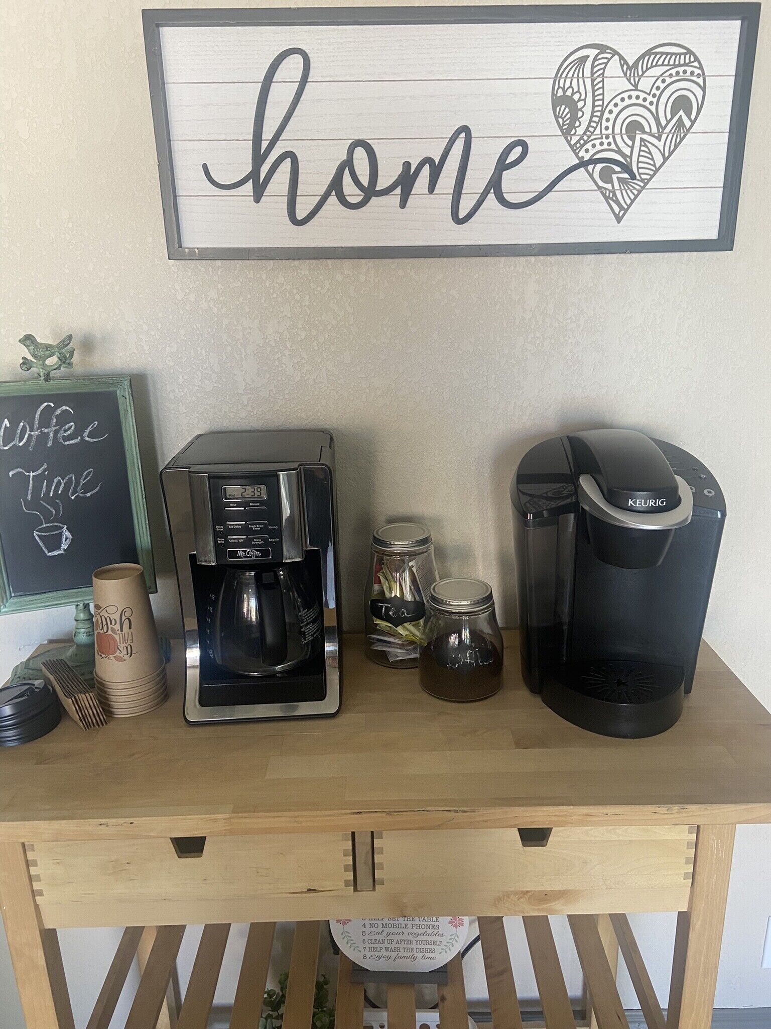 Una estación de café con una máquina Keurig, dos frascos etiquetados como 'té' y 'café', y una pizarra con la inscripción 'hora de café'.