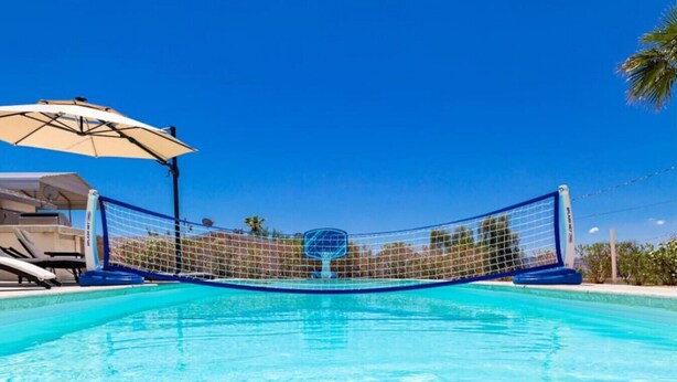 Ein Schwimmbecken mit Volleyballnetz in der Mitte, umgeben von Liegestühlen und einem Sonnenschirm.