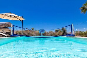 Ein Schwimmbecken mit Volleyballnetz in der Mitte, umgeben von Liegestühlen und einem Sonnenschirm.