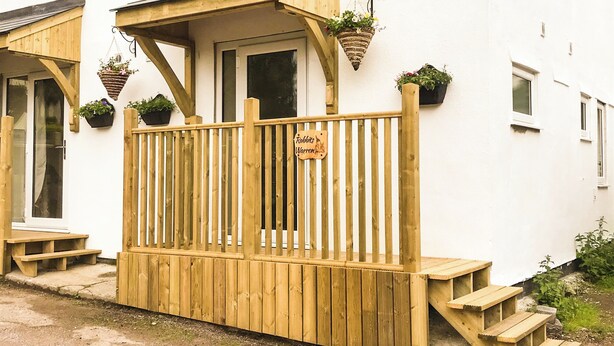 Eine Holzterrasse mit einem Schild, auf dem 'Roberts Warren' steht, einer Tür und Blumentöpfen.