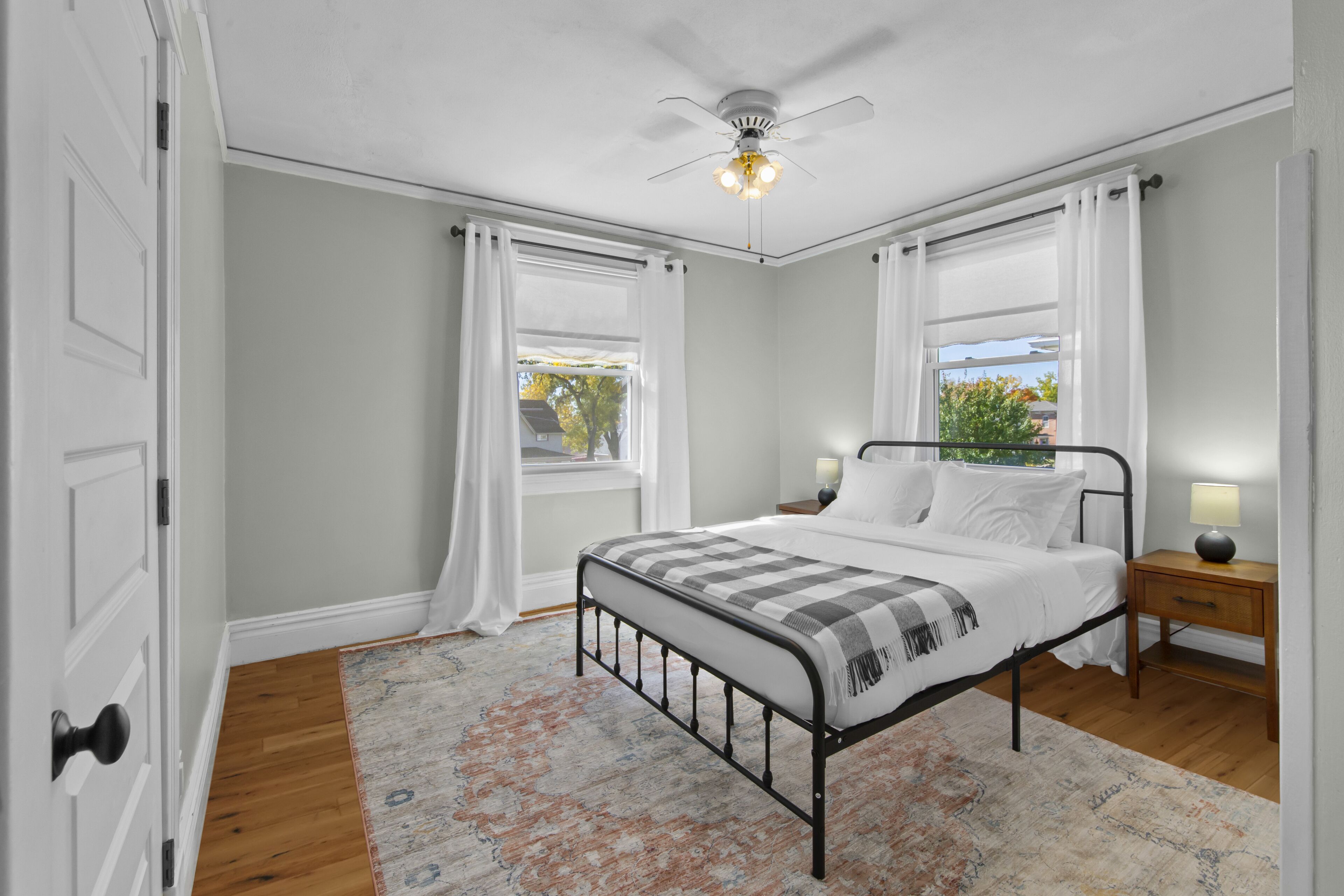 Un dormitorio con cama, mesitas de noche, ventilador de techo y ventanas con cortinas.