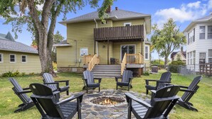 A backyard with a stone fire pit surrounded by Adirondack chairs, a wooden deck, and a house with a chimney.