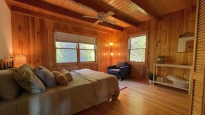 Ein Schlafzimmer mit Holzvertäfelung, einem Bett, zwei Fenstern, einem Sessel, einem Regal und einem Deckenventilator.
