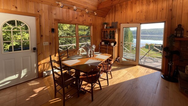 Ein Holzblockhaus-Interieur mit Esstisch, Stühlen, Kamin und großen Fenstern mit Blick auf einen See und Bäume.