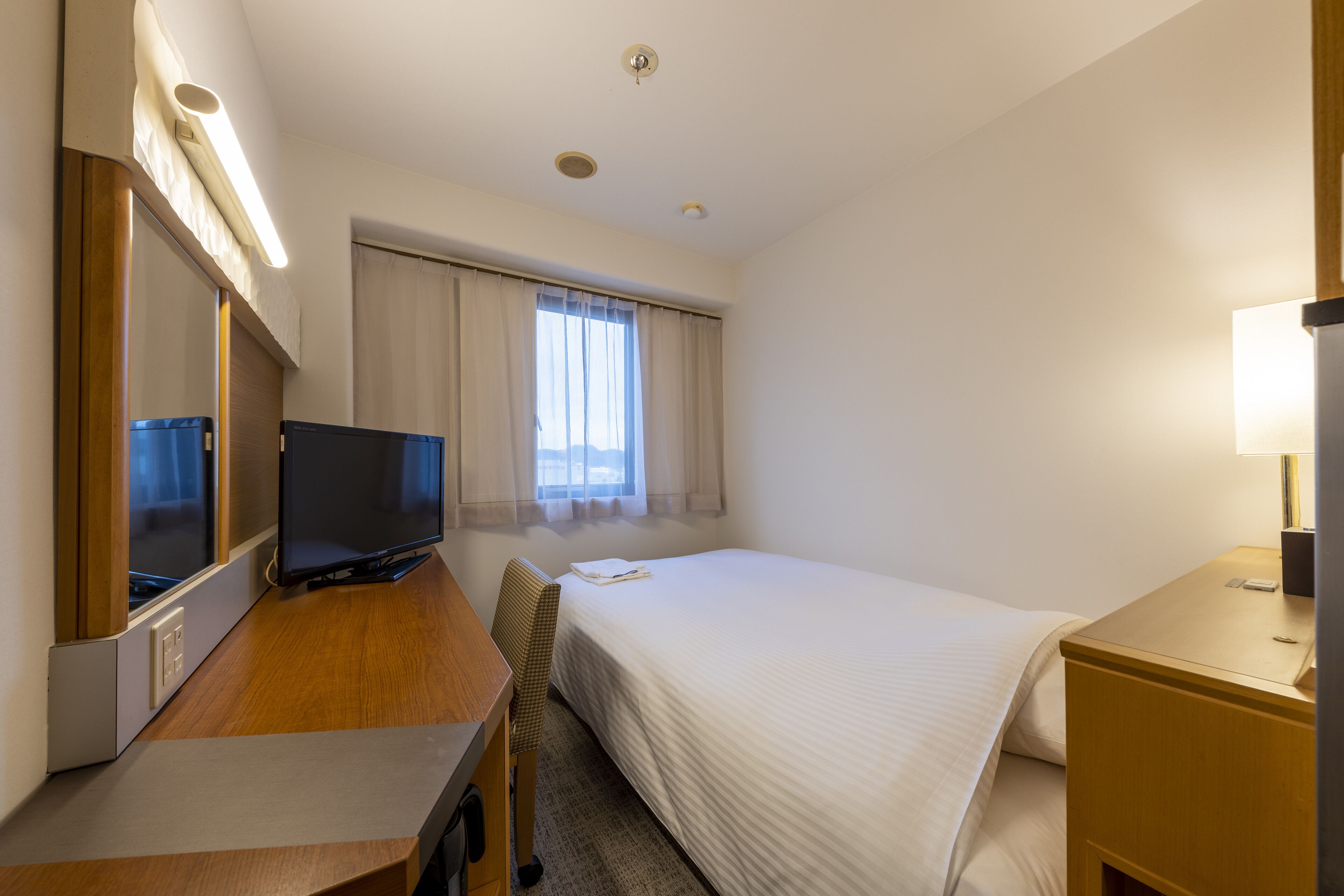Een hotelkamer met een bed, een bureau met een televisie, een stoel, een nachtkastje met een lamp en een raam met gordijnen.