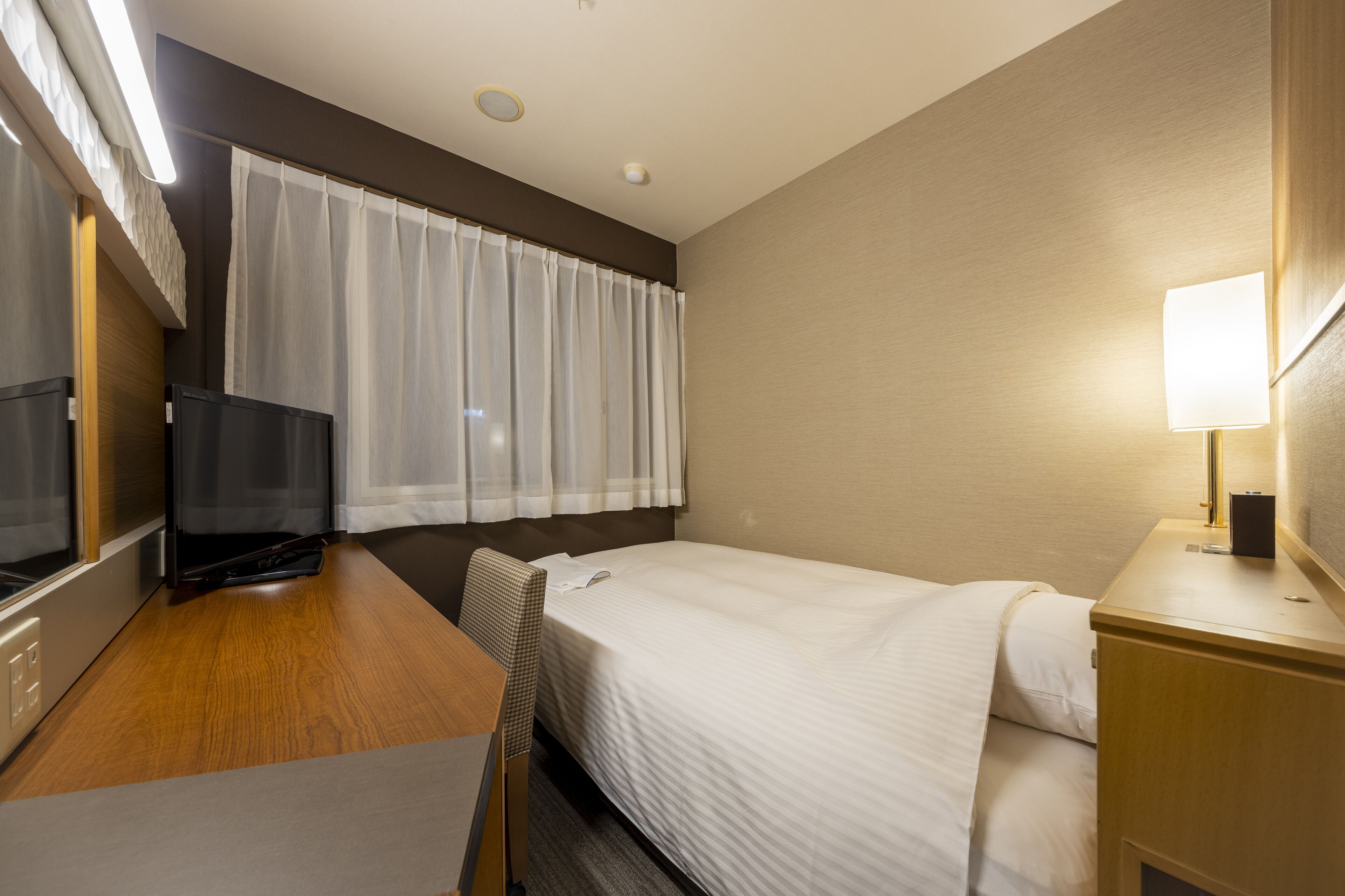 A hotel room with a bed, a desk with a television, a lamp, and a window with curtains.