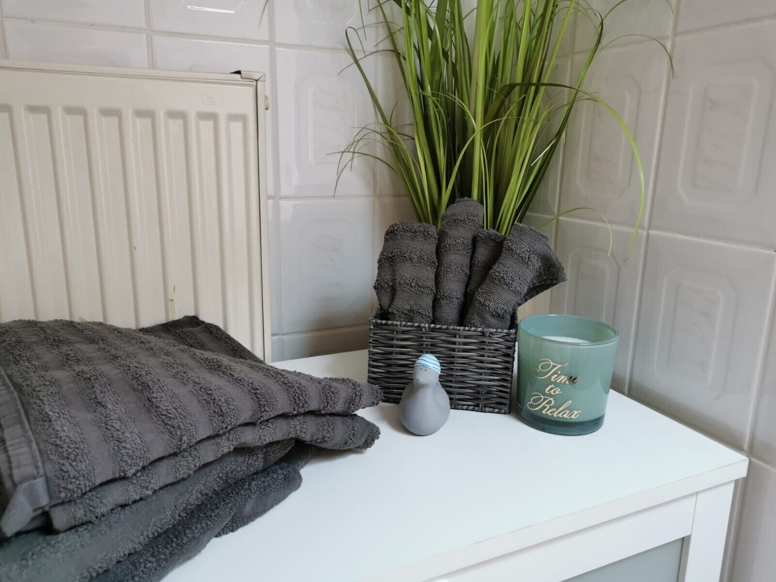 Une salle de bain avec un radiateur blanc, une plante, un panier rempli de serviettes pliées, un vase et une tasse turquoise sur laquelle est inscrit le texte « Il est temps de se détendre ».
