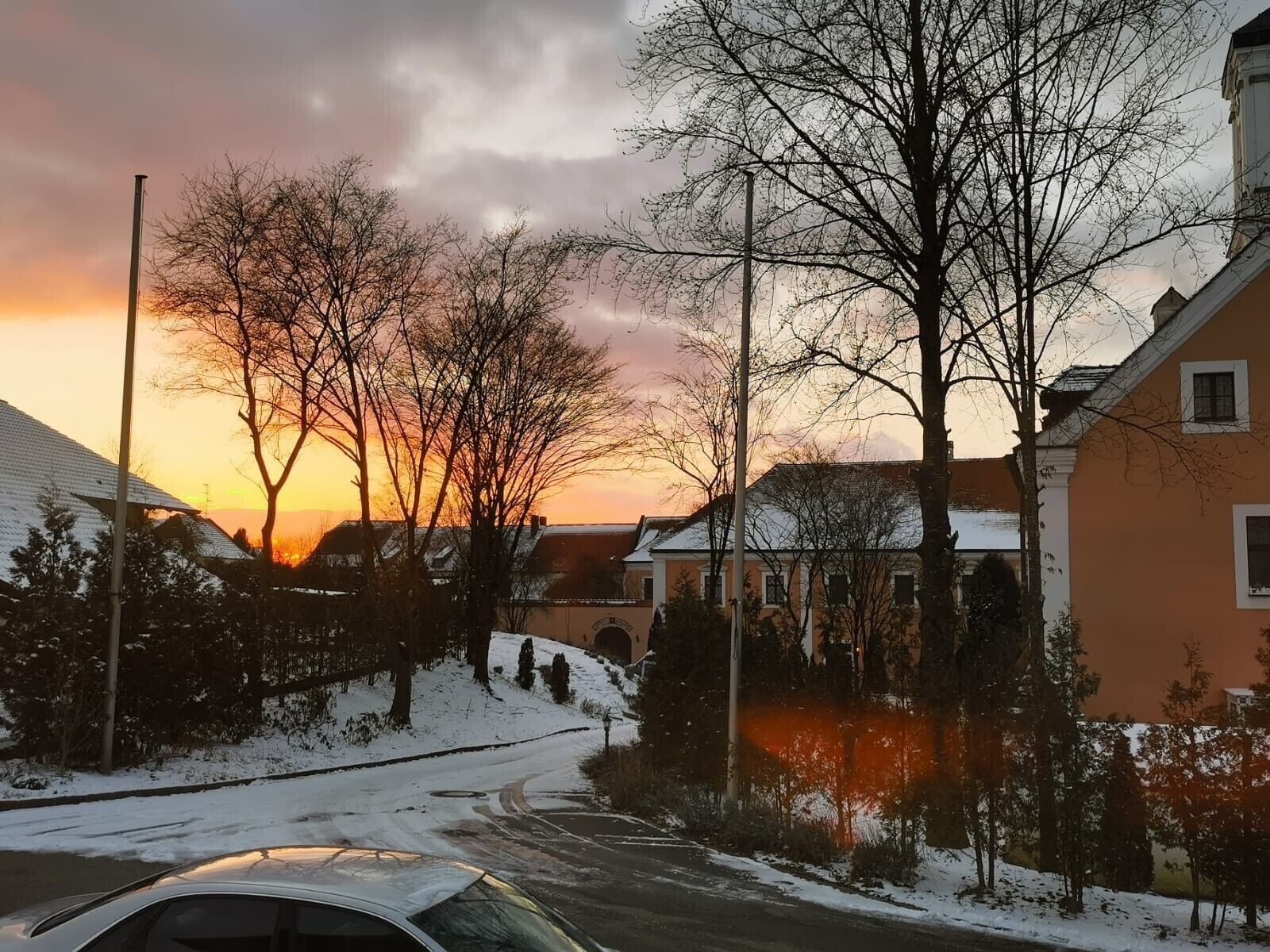 Uma rua residencial com casas, um carro e árvores sem folhas contra um céu de pôr do sol.