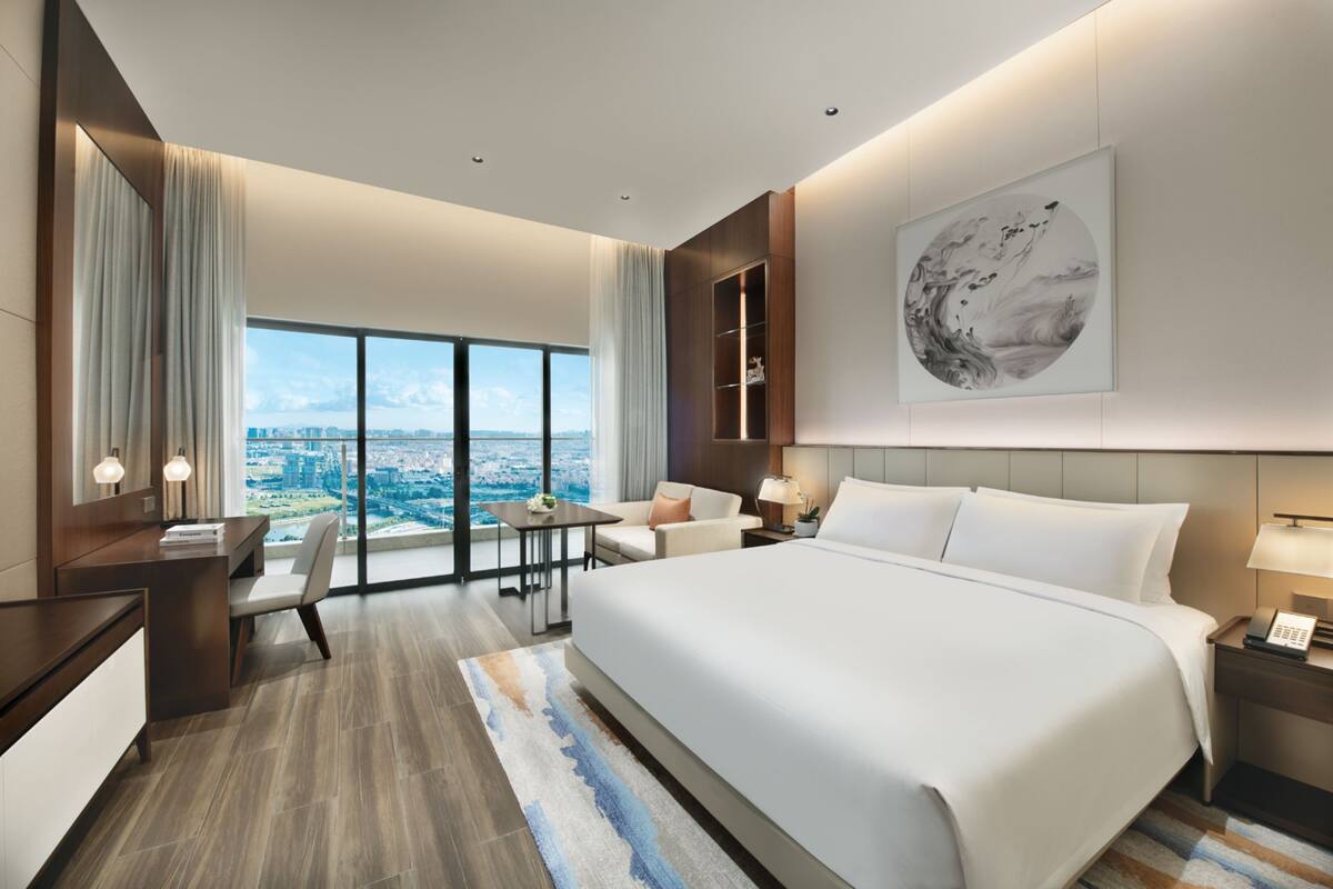 A modern hotel room with a large bed, a desk, a chair, and a view of the city.