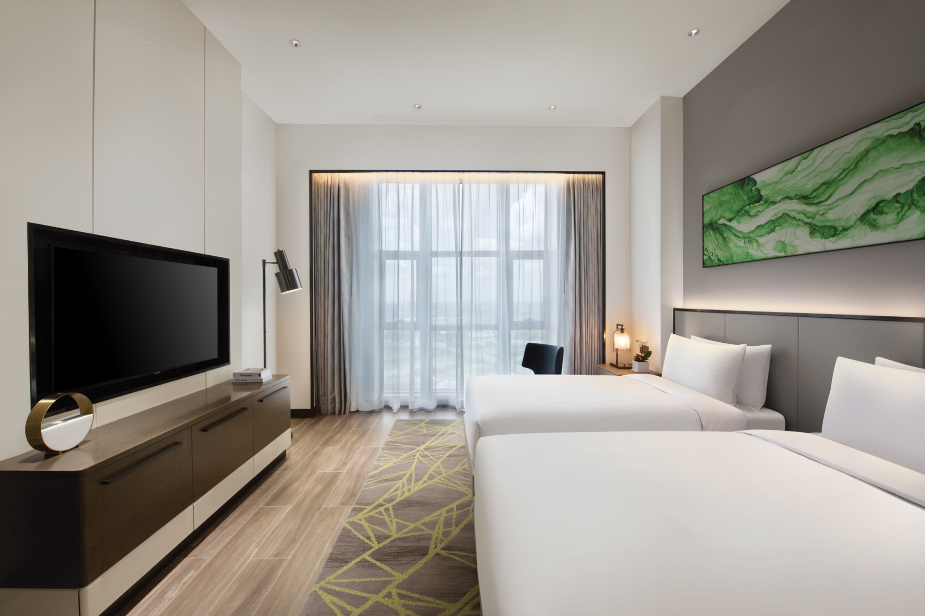 Ett hotellrum med en stor platt-TV, en säng, ett skrivbord med en lampa och ett stort fönster med gardiner.
