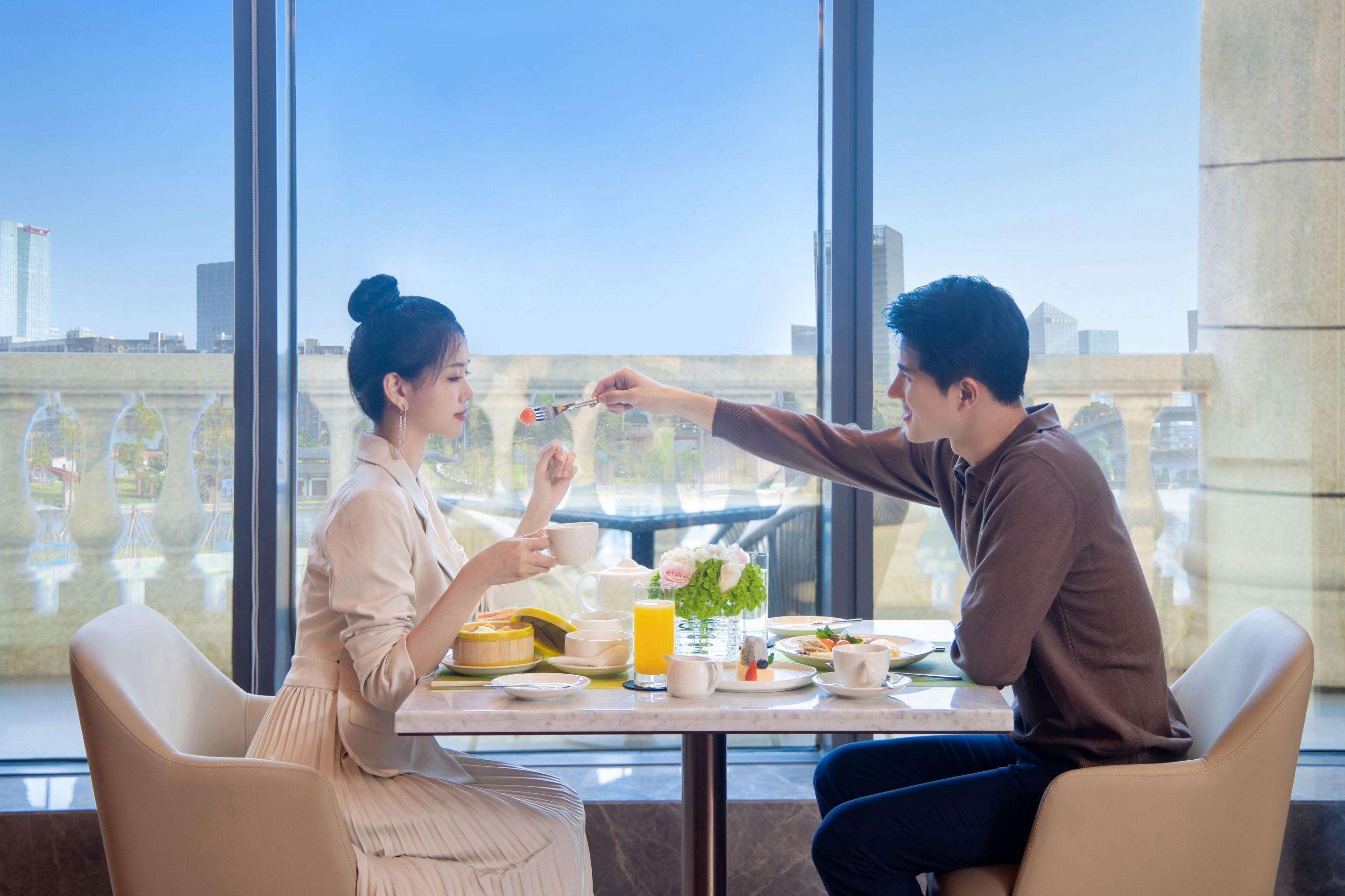 Una mesa de comedor con alimentos para el desayuno, un florero con flores y vistas a la ciudad a través de amplios ventanales.