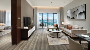 A modern hotel room with a large window offering a city view, a flat-screen TV, a sofa, a coffee table, and a dining area with a table and chairs. - Oakwood Residence Foshan (Foshan)