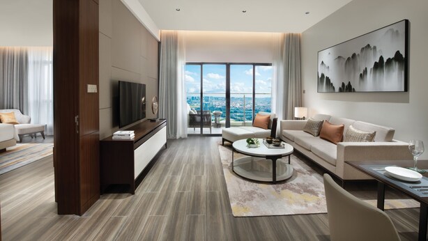 A modern hotel room with a large window offering a city view, a flat-screen TV, a sofa, a coffee table, and a dining area with a table and chairs. - Oakwood Residence Foshan (Foshan)