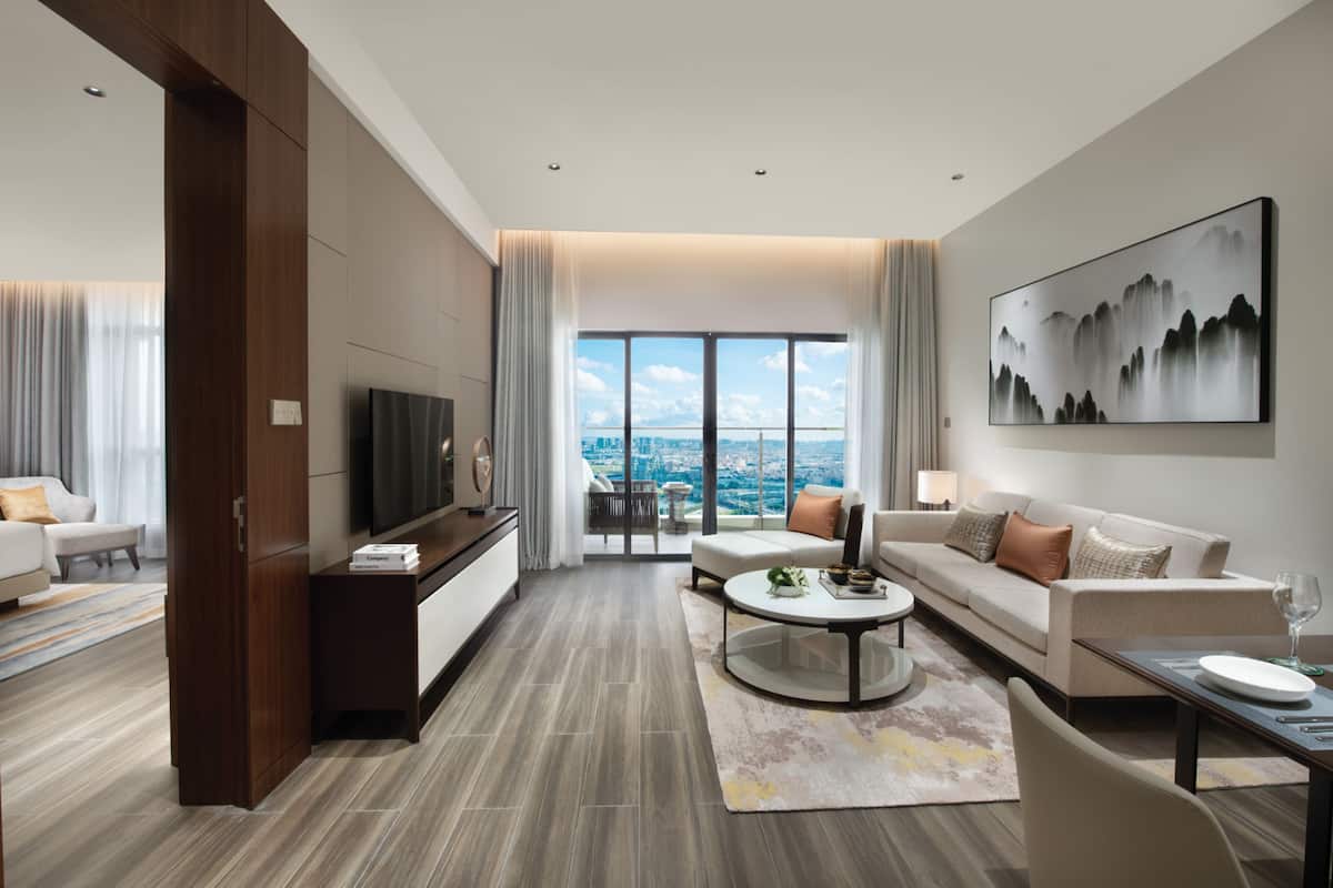 A modern hotel room with a large window offering a city view, a flat-screen TV, a sofa, a coffee table, and a dining area with a table and chairs.