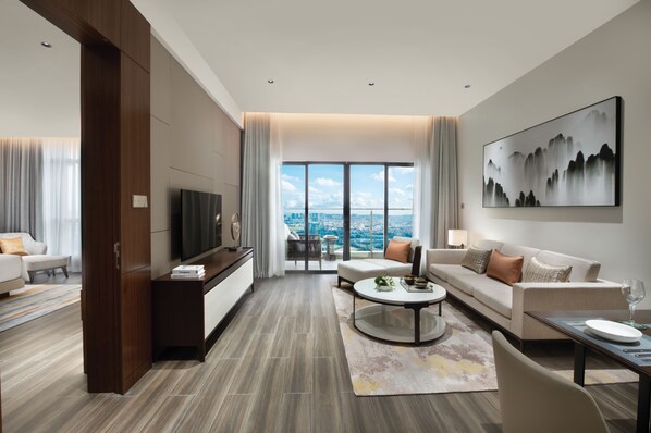 Habitación de hotel moderna con un amplio ventanal que ofrece vistas a la ciudad, un televisor de pantalla plana, un sofá, una mesa de centro y un comedor con mesa y sillas.
