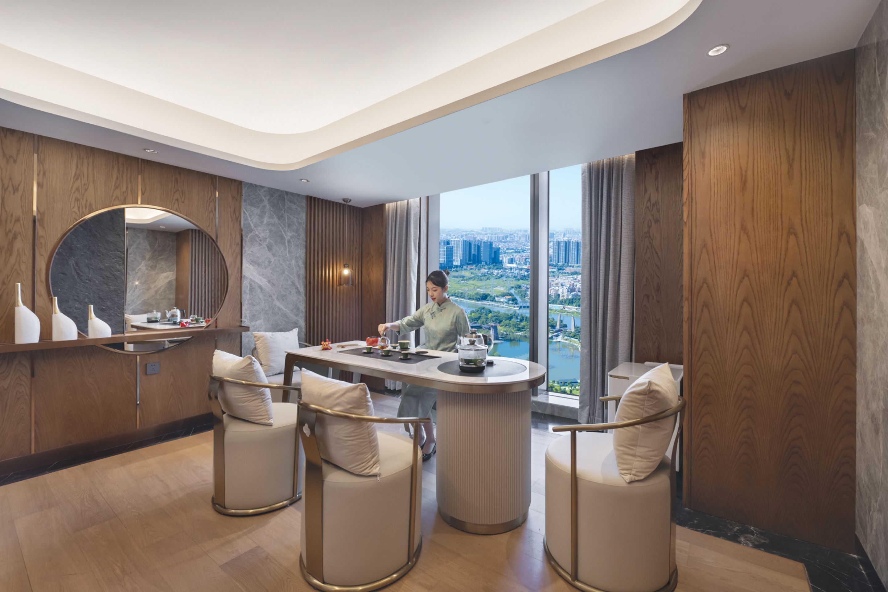 A modern hotel room with a large window offering a city view, a round dining table with chairs, and a bar area.