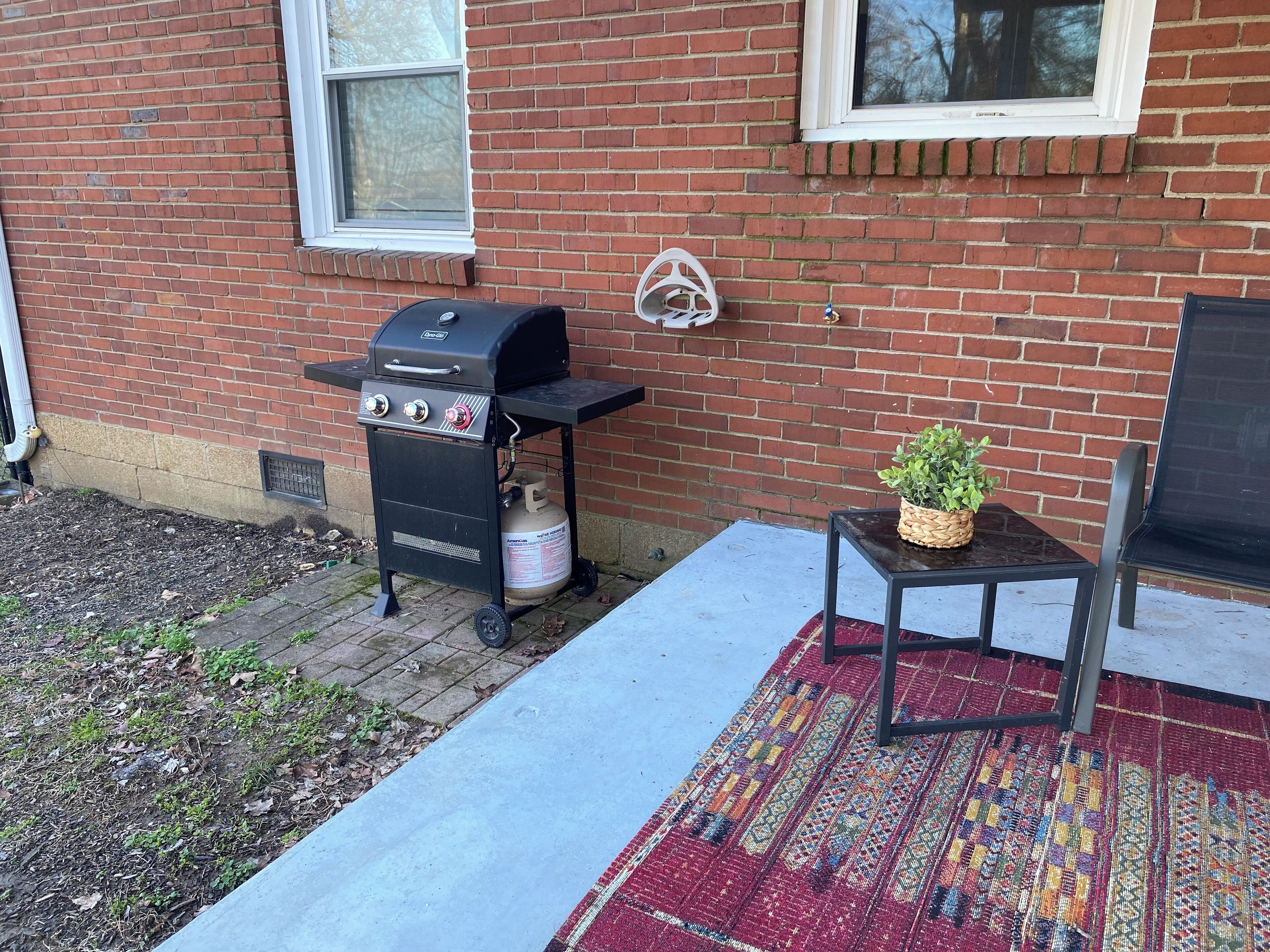 Fachada de una casa de ladrillo con una parrilla de barbacoa y un tanque de gas, una pequeña mesa con una planta, y una alfombra estampada en el porche.