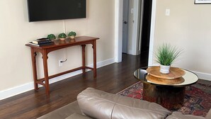 A living room with a beige leather sofa, a wooden console table with books and plants, a flat-screen TV mounted on the wall, and a round coffee table with a plant. - Comfortable and Quiet 3 Bedroom Duplex (Nashville)