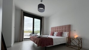 A modern bedroom with a large bed, a bedside table with a lamp, and a view through a large window. - Atico apartment with views of the Casares golf course, 3,00 metres from Finca Cortesin (Casares Costa Golf)