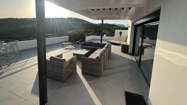 A modern outdoor patio with wicker furniture, a glass table, and a view of a hilly landscape. - Atico apartment with views of the Casares golf course, 3,00 metres from Finca Cortesin (Casares Costa Golf)