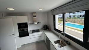 A modern kitchen with white cabinetry, a built-in oven, a stainless steel sink, and a window overlooking a swimming pool. - Atico apartment with views of the Casares golf course, 3,00 metres from Finca Cortesin (Casares Costa Golf)