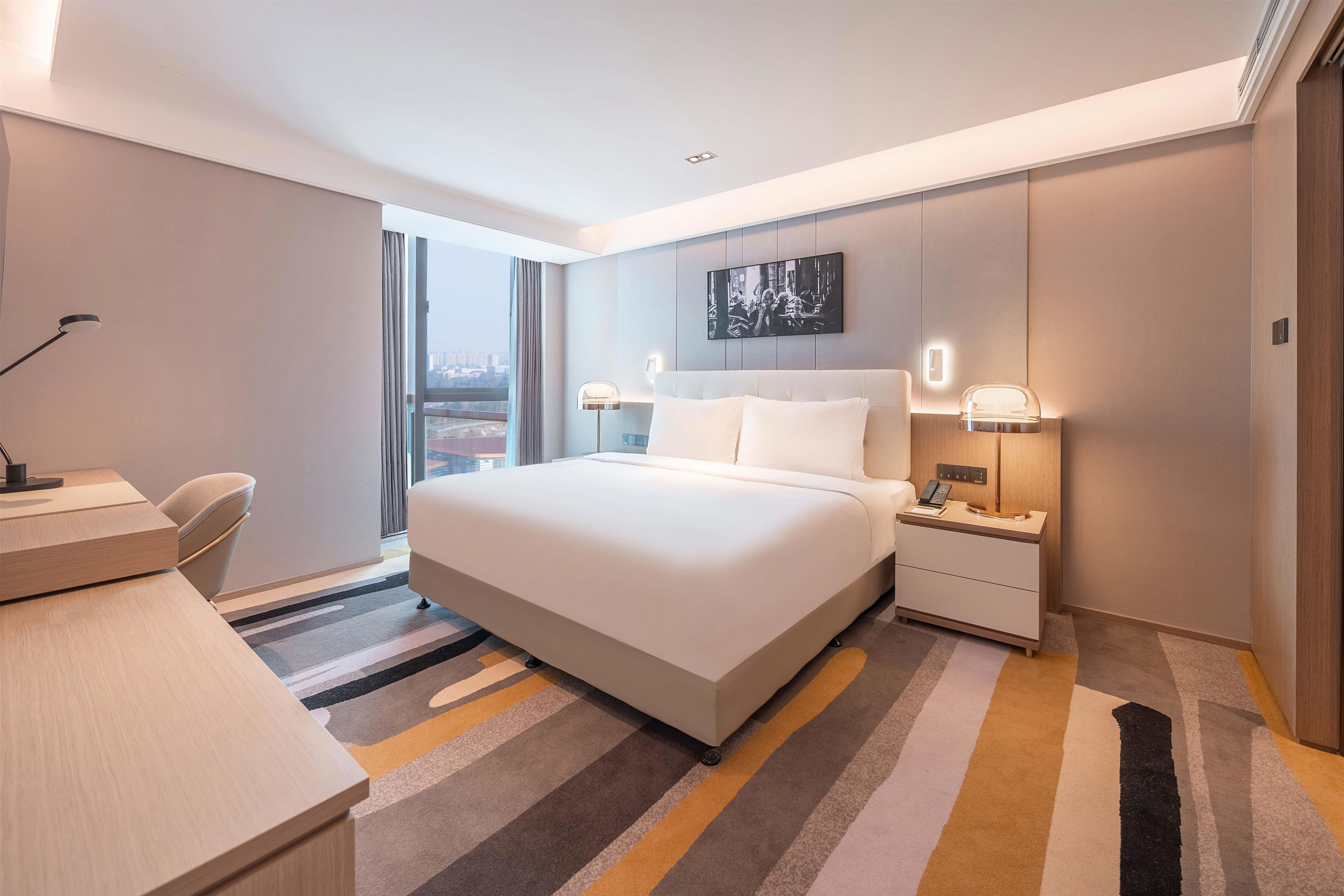 A modern hotel room with a large bed, bedside lamps, a desk, and a view of the city.