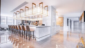 A modern hotel lobby with a marble reception desk, a bar area with stools, and a seating area with chairs.