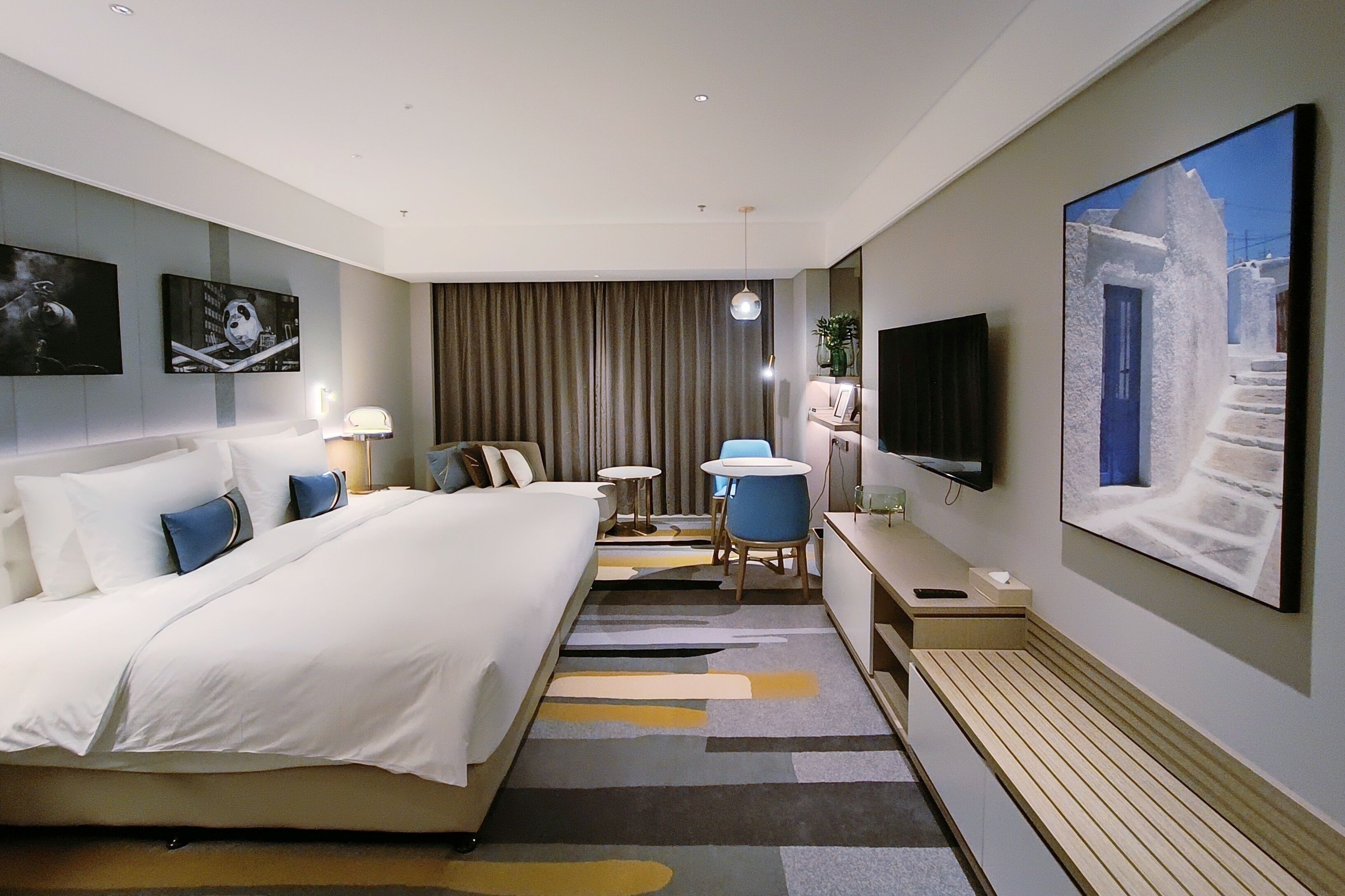 Une chambre d’hôtel avec un grand lit, un bureau avec une chaise, une télévision et des œuvres d’art accrochées aux murs.