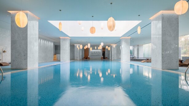 A spacious indoor swimming pool with a clear blue water surface, surrounded by marble columns and modern pendant lighting.