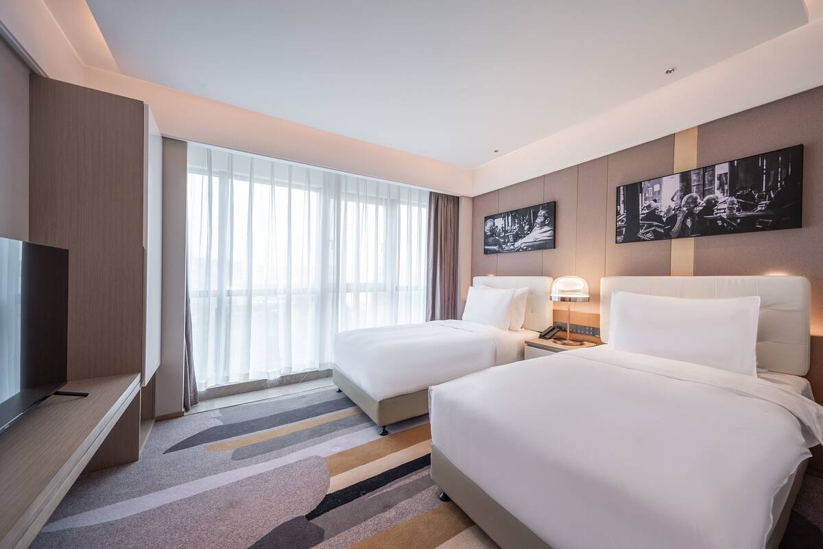 Une chambre d’hôtel avec deux lits, une télévision, un bureau et une grande fenêtre avec des rideaux.