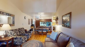 A living room with a sofa, a patterned armchair, a coffee table, and a rug. There is a dining area with a table and chairs, and a kitchen in the background. - Pet Friendly | Close to Lifts | Fireplace | Charming | Angel Fire Chalet 52 (Angel Fire)
