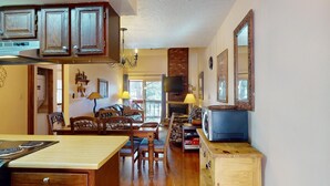 A cozy kitchen with wooden cabinets, a dining area with chairs, a TV mounted above the fireplace, and a wooden cabinet with a microwave. - Pet Friendly | Close to Lifts | Fireplace | Charming | Angel Fire Chalet 52 (Angel Fire)