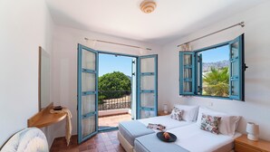 A bedroom with a bed, bedside tables, a desk, and a balcony with a view of greenery and mountains. - Villa Oceanis (Lindos)
