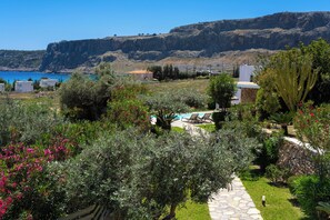 A coastal property with a stone pathway leading to a pool area, surrounded by lush greenery and overlooking a cliff and the sea.