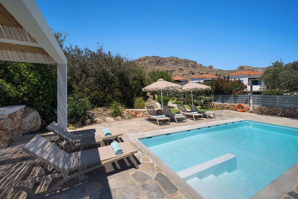 A swimming pool with lounge chairs and umbrellas, surrounded by greenery and a mountainous backdrop.
