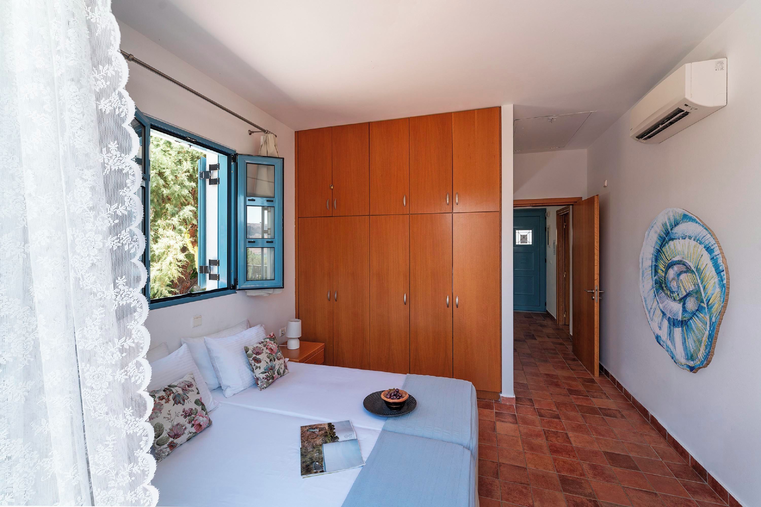 A bedroom with a bed, a wardrobe, a window with blue shutters, and a wall-mounted air conditioner.