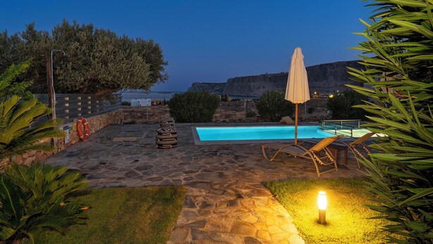 A stone patio with a pool, lounge chairs, and a well-lit pathway. - Villa Oceanis (Lindos)