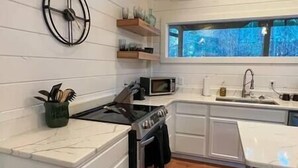 A kitchen with white cabinetry, a stove, a microwave, a sink, and a window with a view of trees. - SERENITY NOW · Relax & Unwind (Broken Bow)