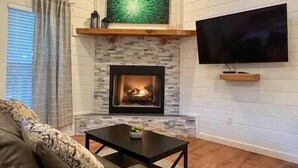 A living room with a fireplace, a television mounted on the wall, and a window with curtains. - SERENITY NOW · Relax & Unwind (Broken Bow)
