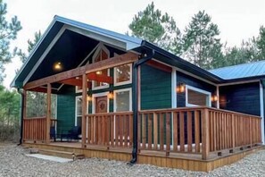 A cabin with a wooden deck, a covered porch, and a large window. - SERENITY NOW · Relax & Unwind (Broken Bow)