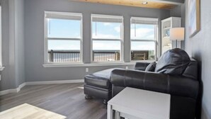 A modern living room with a gray wall, a black leather sofa, a white side table, and a view of the ocean through large windows. - Stunning Beachfront 3 Bedroom Home, CoralMoonBeach (Gilchrist)