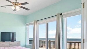 A bedroom with a bed, a TV, a ceiling fan, and a view of the beach through large windows. - Stunning Beachfront 3 Bedroom Home, CoralMoonBeach (Gilchrist)
