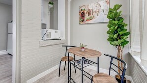 Un petit coin repas avec un mur en briques blanches, un tableau représentant une scène de rue et une grande plante verte.