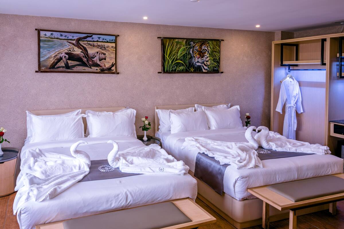 A hotel room with two beds, each with white linens and decorative swan-shaped towels. There are paintings on the walls, a small table with a vase of flowers, and a robe hanging on a rack.